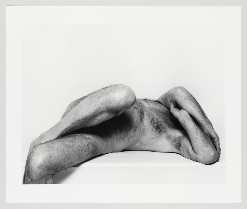A hairy nude male reclining aganist a flat surface with crossed arms and legs