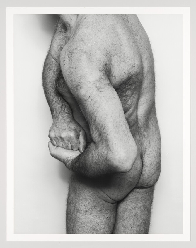 Adult man standing in profile with hands clasped behind his back, showing bare torso and buttocks.