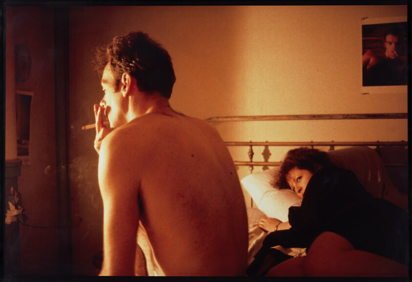 Shirtless man sits on a bed smoking a cigarette while a woman lies behind him watching.