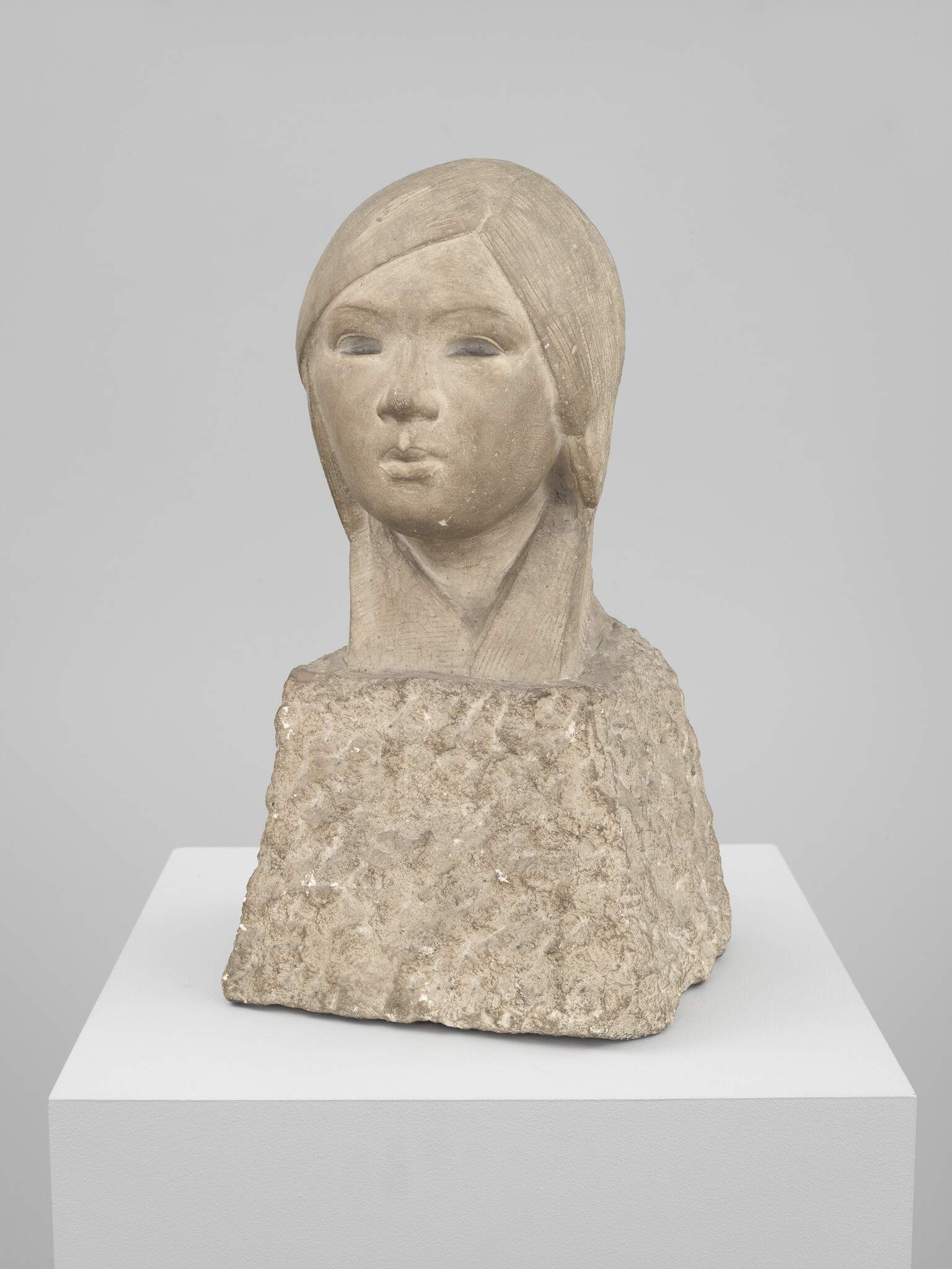 Stone bust of a young girl with stylized hair mounted on a rough rectangular base.