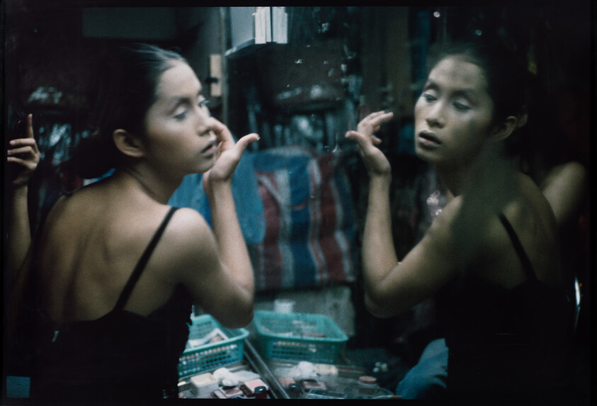 Young woman applies makeup at a cluttered vanity, her reflection looking back.