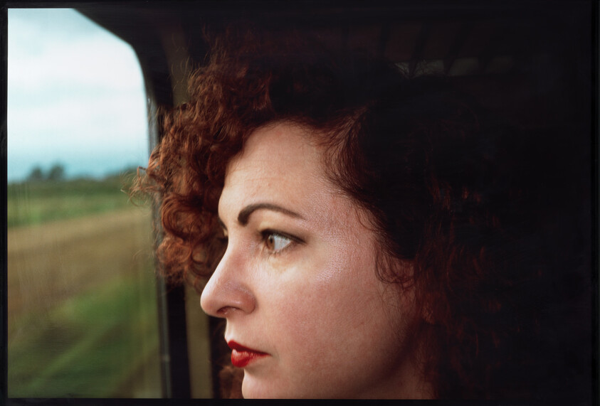Woman with curly hair looks pensively out a window at passing fields.