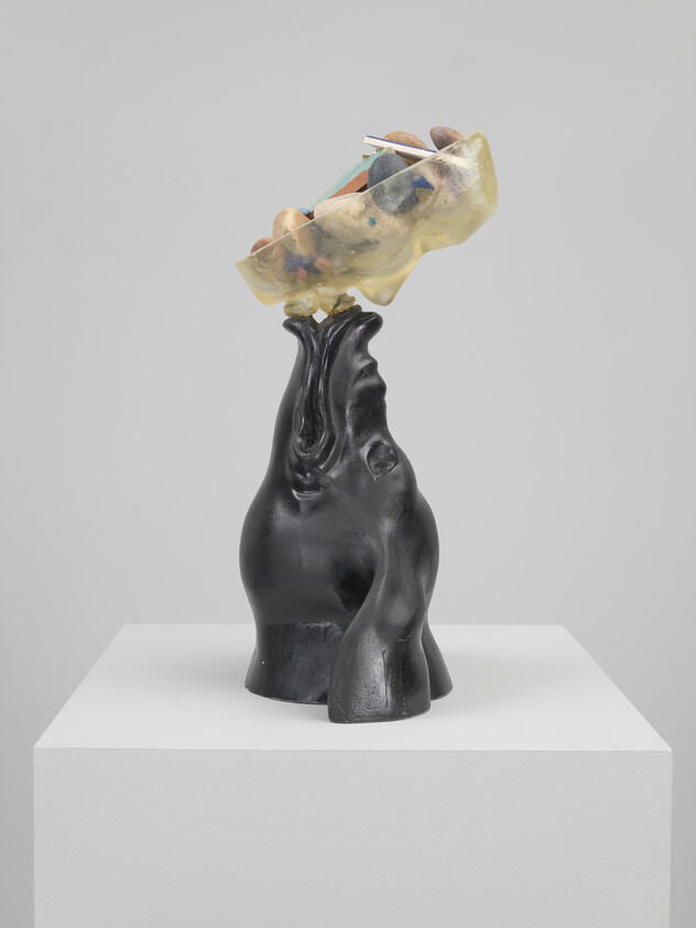 A sculpture of a stylized black torso with an abstract, multicolored form on top, resembling a head or a hat, displayed on a white pedestal against a light grey background.