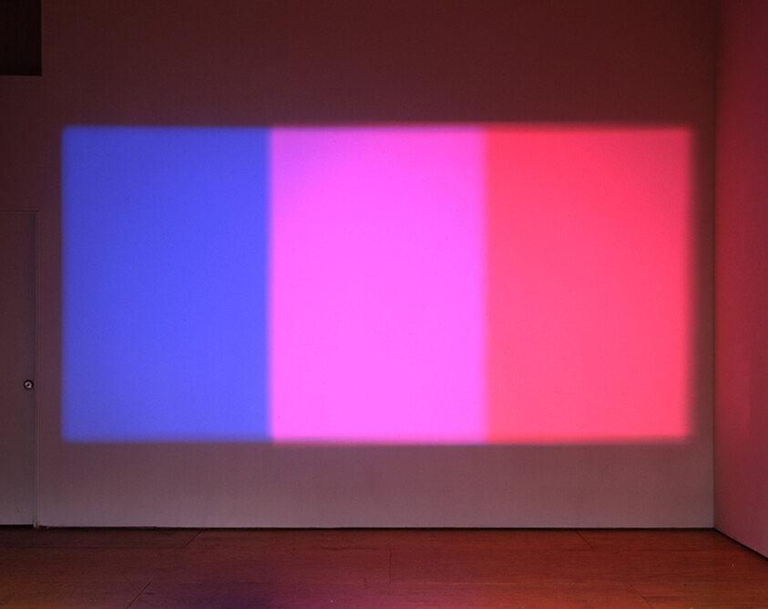 Three colored light projections on a wall: blue, purple, and red, blending where they meet.