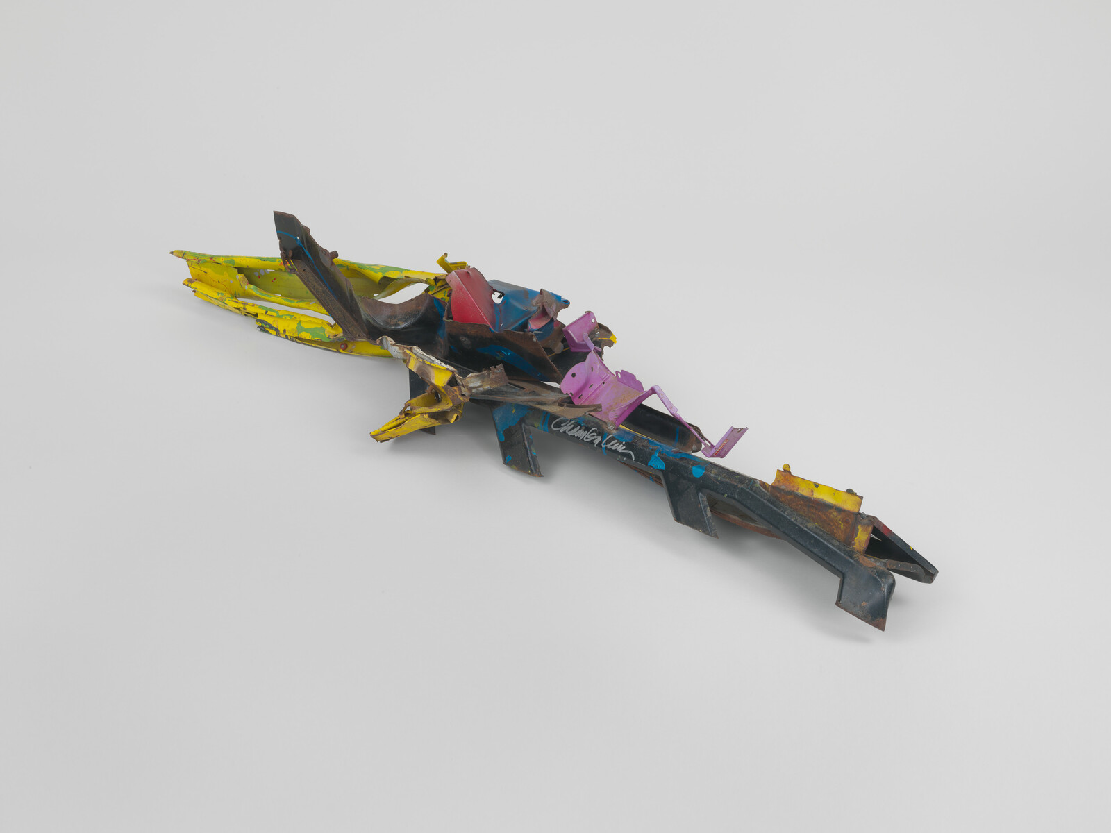 Long twisted scrap metal sculpture painted yellow, blue, pink, and black resting on a white surface.