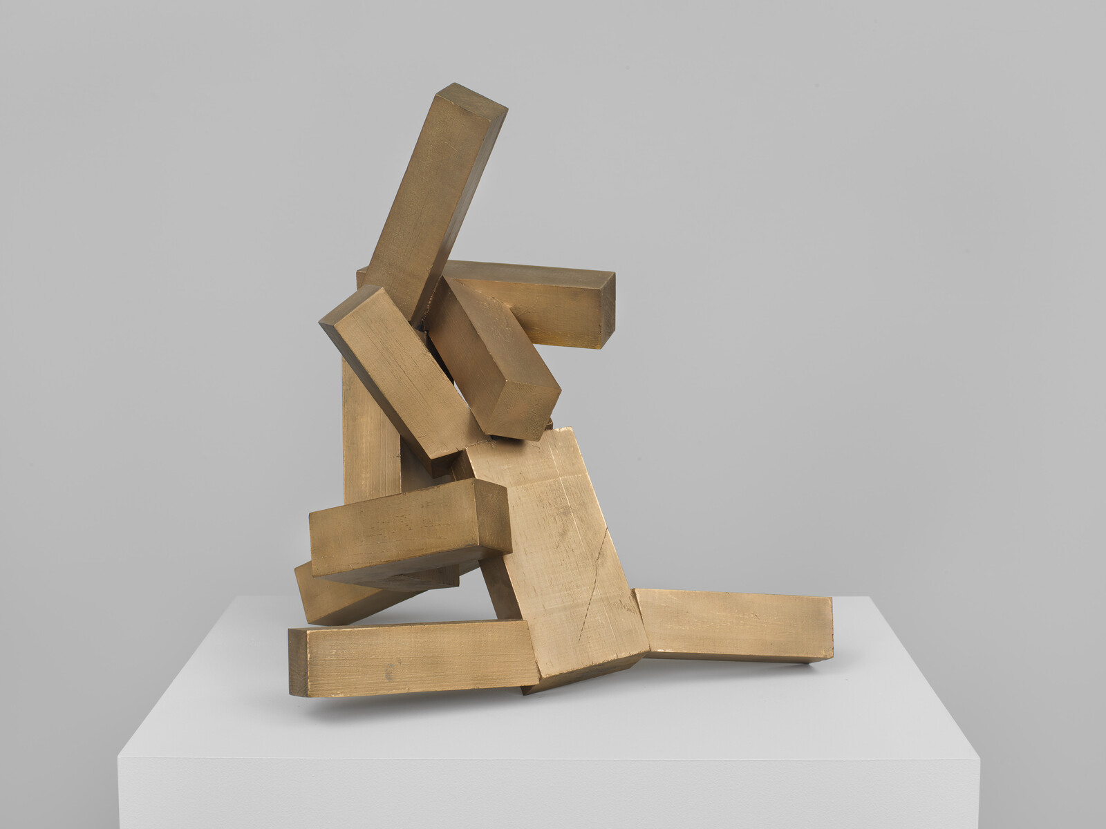 Stacked bronze rectangular blocks arranged into an abstract leaning sculpture on a white pedestal.