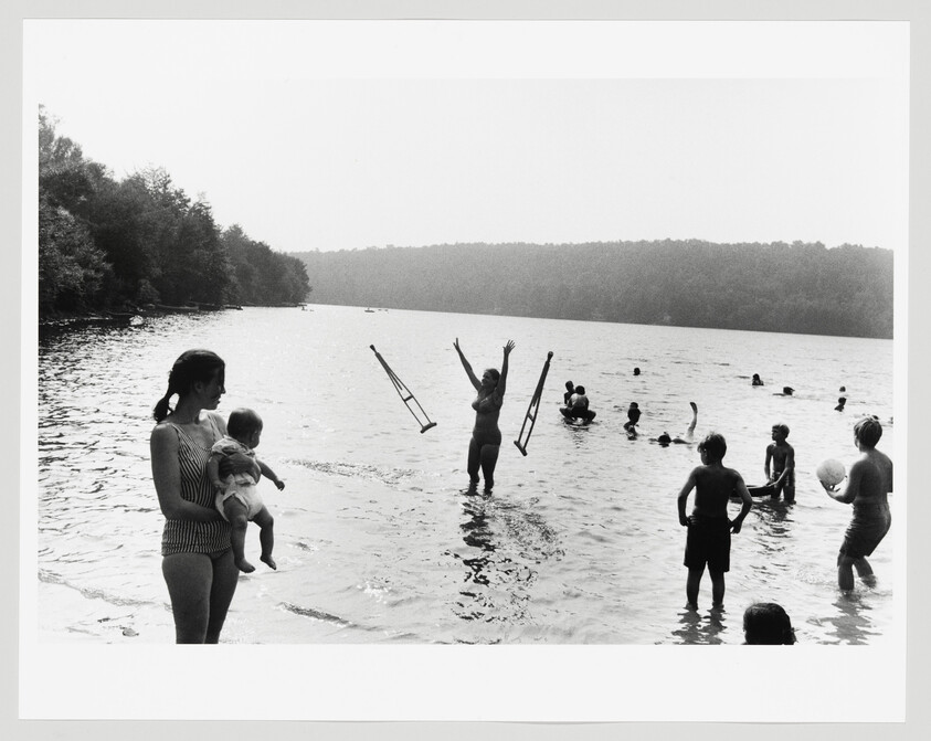 Several people swim and play in a lake while a woman tosses crutches into the air.