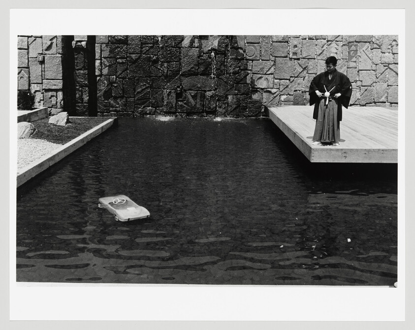 A person in traditional Japanese clothing stands on a platform watching a small toy car float in the pond.