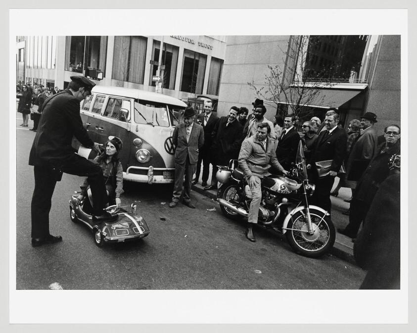 Policeman leans toward woman sitting in toy car while crowd watches near motorcycle and van.