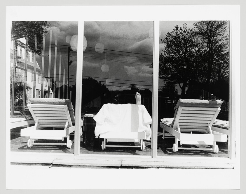 Three lounge chairs behind a glass window, a covered person on the middle chair with one foot raised.
