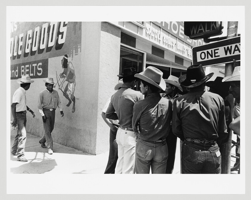 A group of men wearing cowboy hats stand and talk on a city sidewalk beneath a one-way sign.