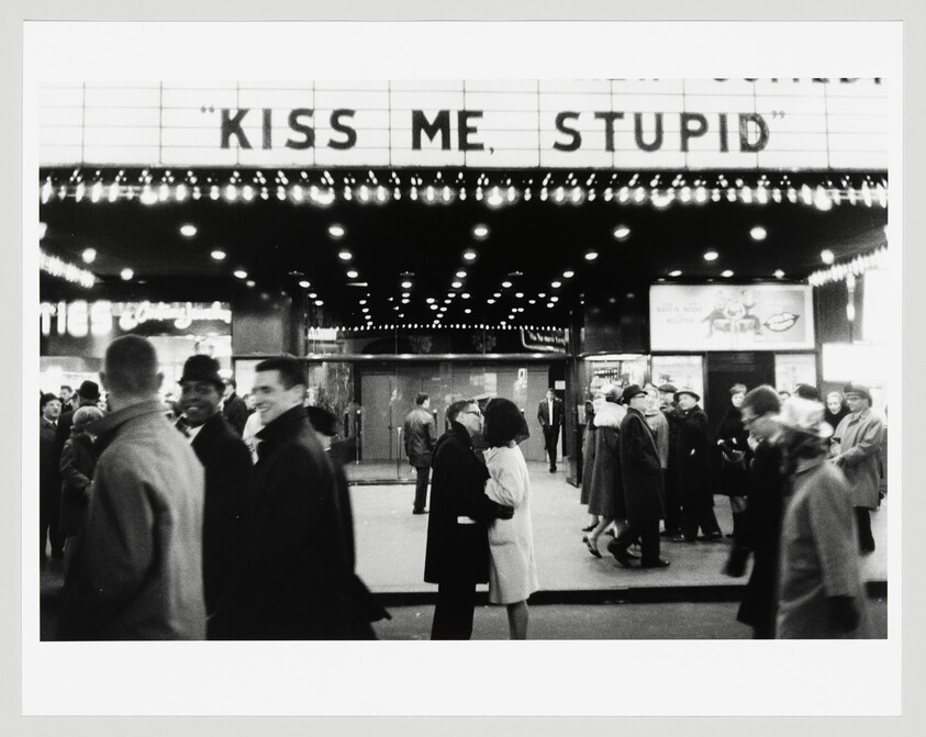 A black and white photograph of a bustling movie theater entrance with the marquee reading "KISS ME, STUPID" in large letters. People are seen mingling and walking around the entrance, with some in the foreground appearing blurred due to motion, suggesting the liveliness of the scene. The theater's facade is adorned with a string of illuminated bulbs, adding to the classic cinematic ambiance.
