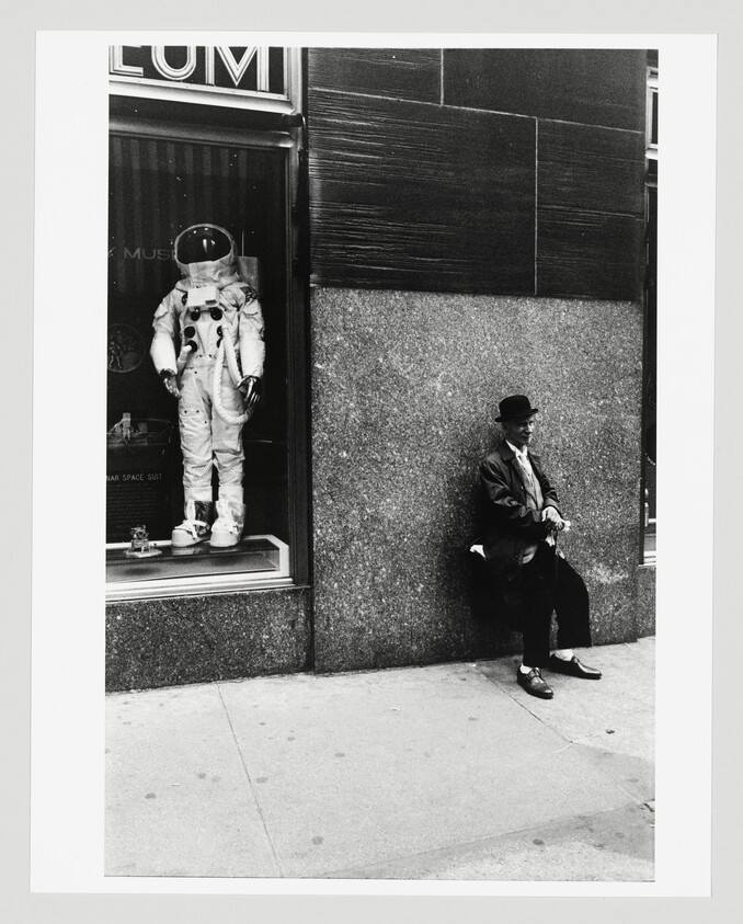 A suited astronaut display stands in a window while a man in a hat sits against the wall.