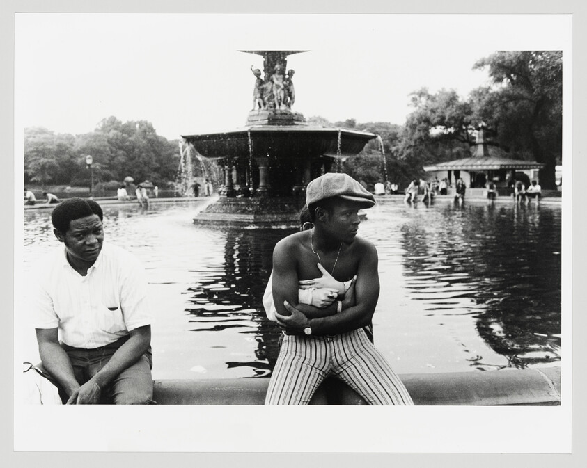 Black and white photograph of two young men sitting by a fountain pool, one looking away thoughtfully and the other gazing into the distance, with the fountain and park visitors in the background.