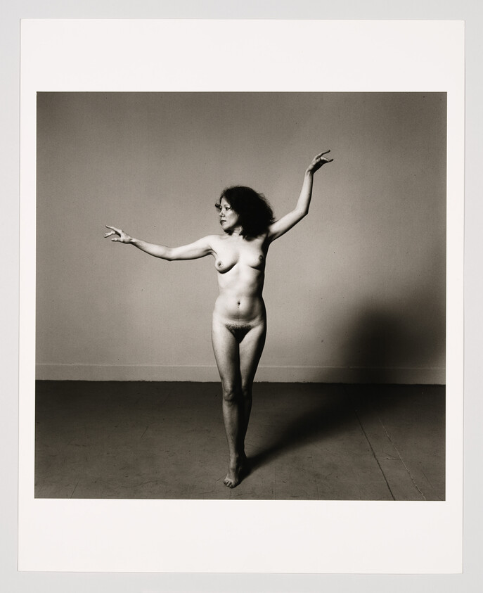 A nude woman standing on one foot with arms raised, looking to her left.