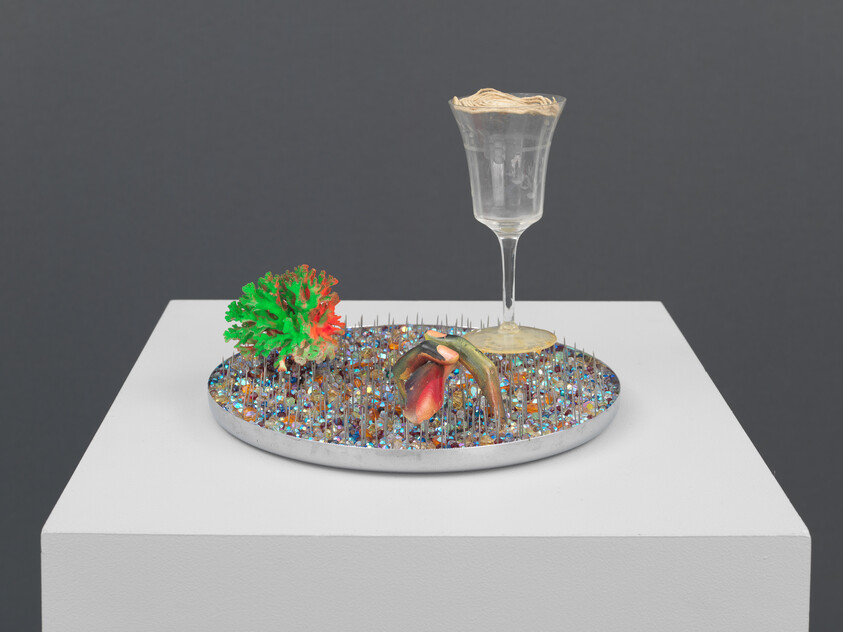 A glass with a beige substance sits on a spiked, colorful base with a plant and shell.