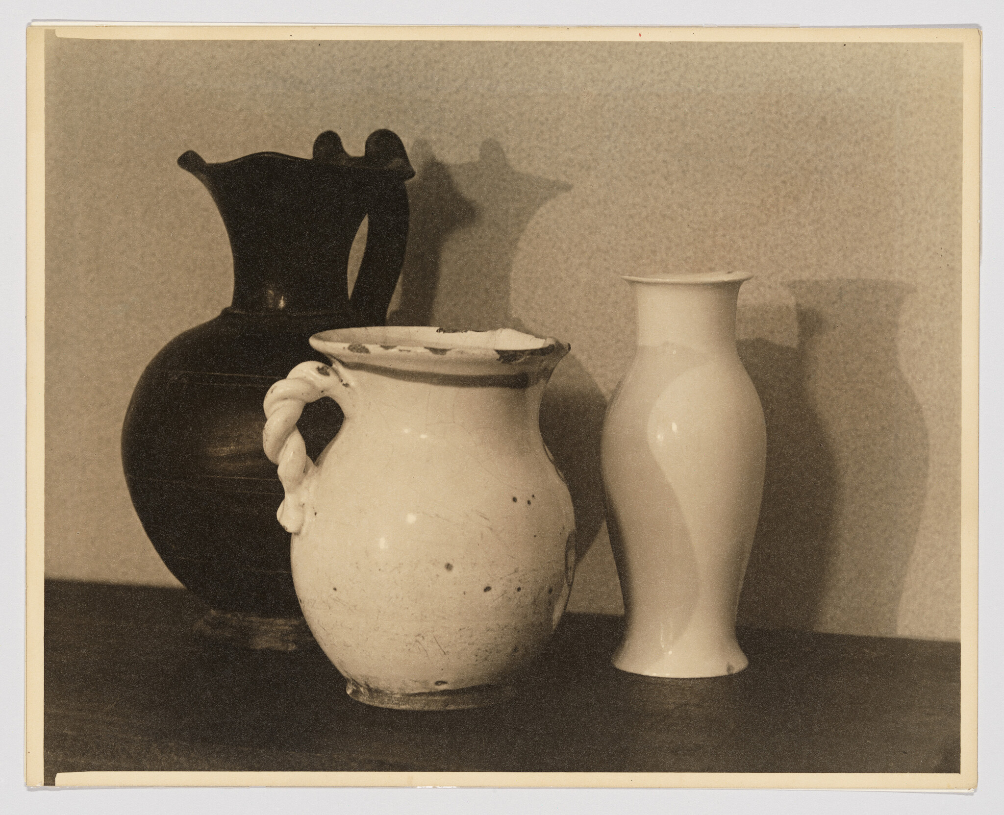 A chipped white jug with a twisted handle sits between a dark pitcher and a tall white vase.