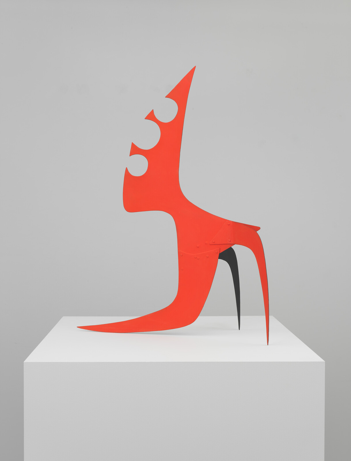 A bright red abstract metal sculpture with sharp pointed legs and crescent cutouts sits on a white pedestal.