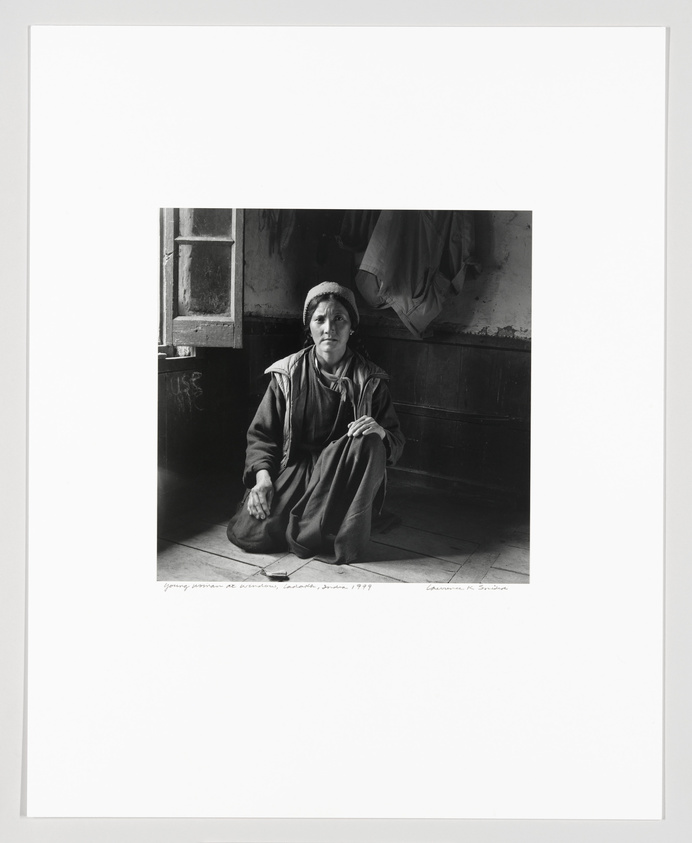 Woman in traditional attire sitting on the floor of a rustic room, looking contemplatively to the side.