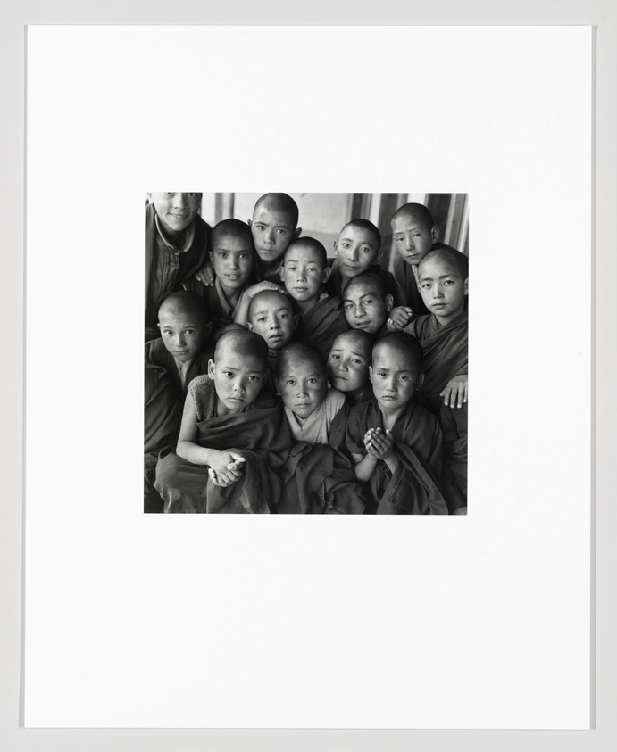 A black and white photograph of a group of young Buddhist monks with shaved heads, wearing traditional robes, gathered closely together and looking directly at the camera.