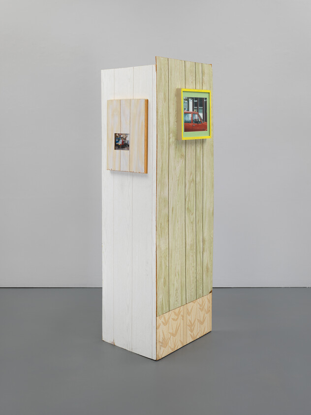 Tall wooden pedestal with pale planks displaying two small framed photographs, one in a bright yellow frame.