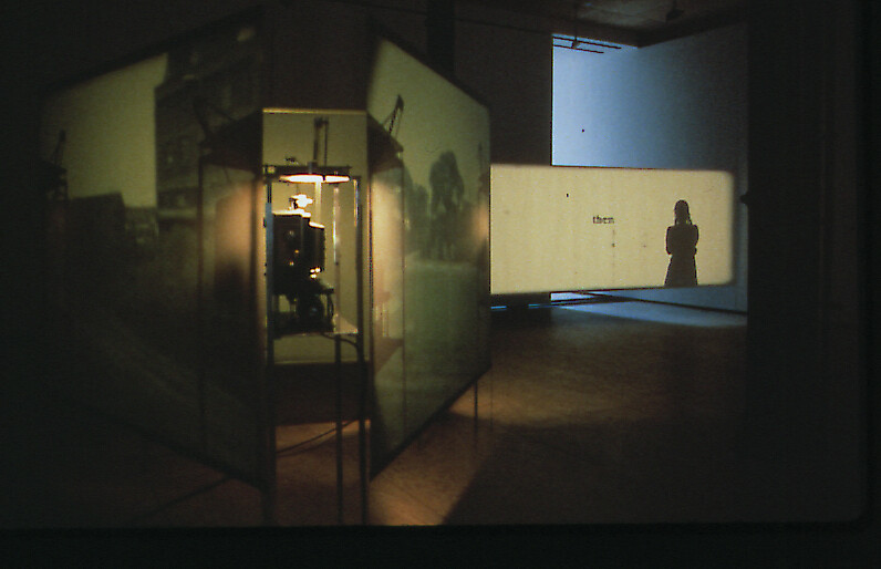 A dark room with projected images on screens, a person’s shadow, and the word “them” visible on one screen.