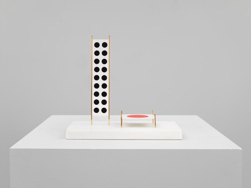 Minimalist sculpture with vertical panel of black circles and small platform featuring a red circle on a white base.