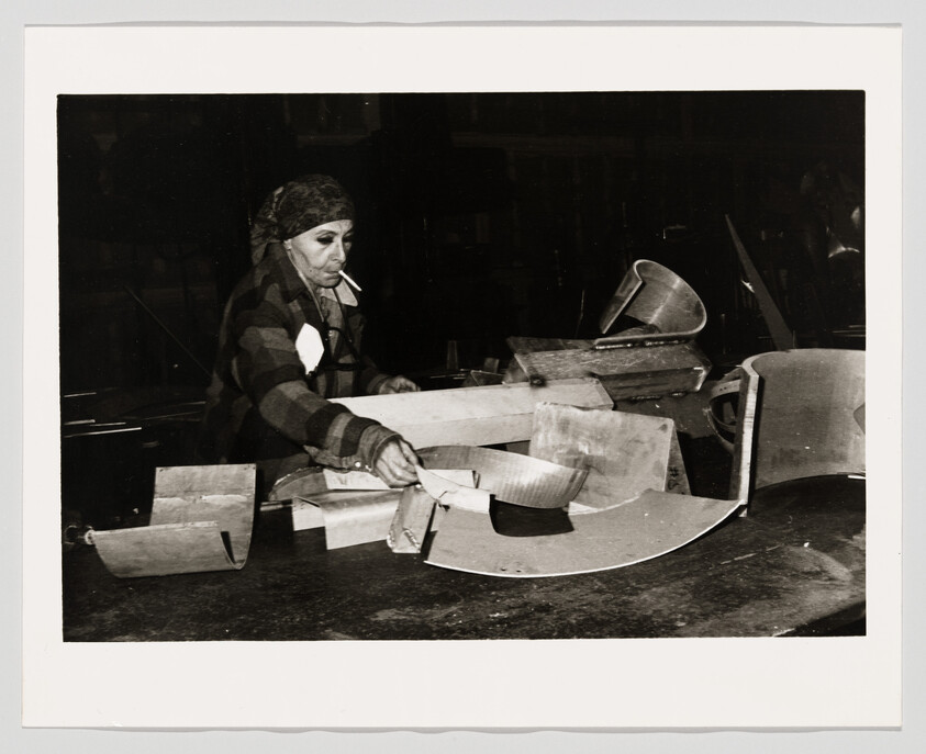 Older woman wearing a headscarf assembles metal parts on a workbench while smoking a cigarette.