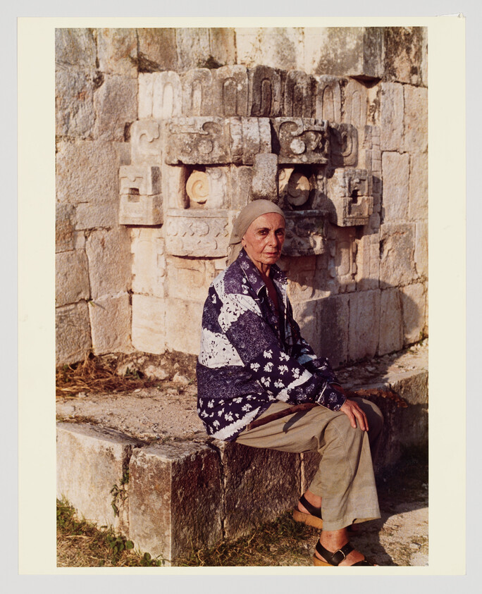 An elderly woman sits on a stone ledge in front of carved ancient stonework.