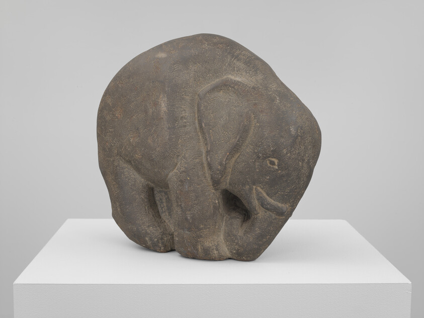 Small carved stone elephant sculpture standing on a white display pedestal in a gallery.