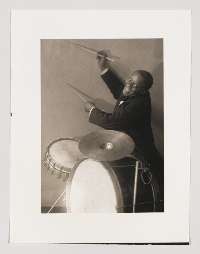 A smiling drummer in a suit energetically raises drumsticks above a bass drum and cymbal.