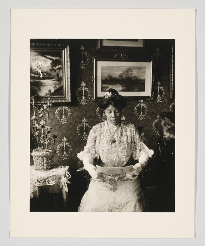 A woman in a lace dress and gloves sits in a decorated parlor reading a letter.