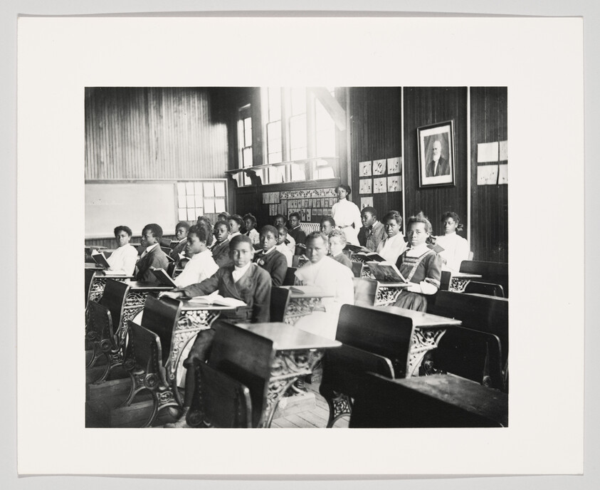 African American students sit at wooden desks in a classroom while a teacher stands at the back.