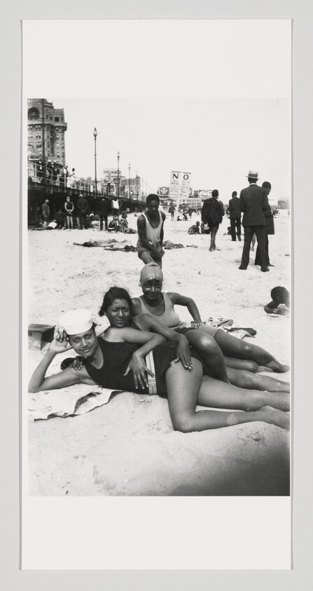 Three women in swimsuits relax and pose together on a sandy beach near a busy boardwalk.
