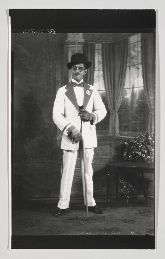 Well-dressed man in a white suit and bowler hat stands holding a cane in a studio.