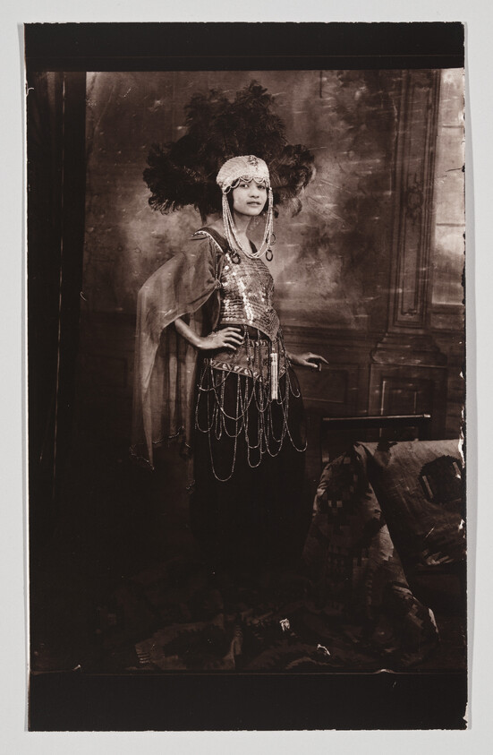 A woman wearing a feathered headdress and beaded costume stands with one hand on her hip.