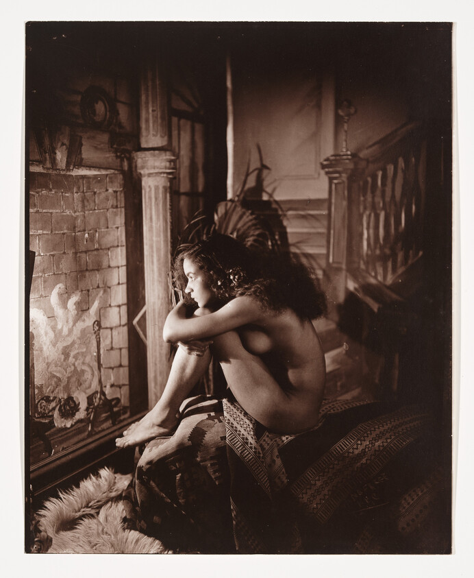 A nude woman sits curled by a lit fireplace, gazing thoughtfully into the flames.