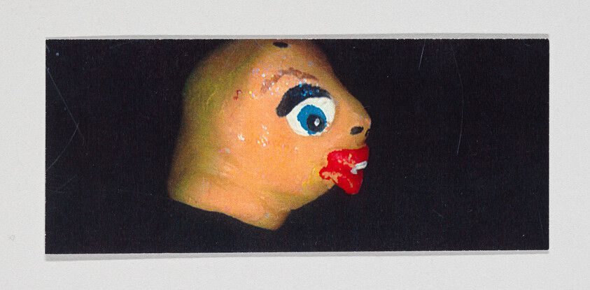 A close-up photograph of a painted object resembling a cartoonish face with one large blue eye, exaggerated red lips, and a red smudge below the lips on a black background.