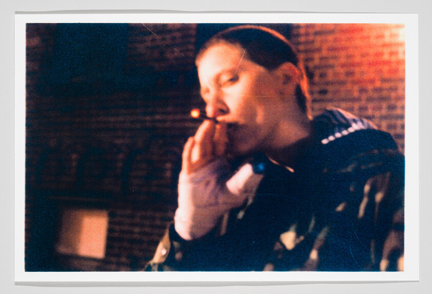 A blurry photograph of a person lighting a cigarette, with a dark, brick wall in the background. The image has a vintage feel with warm tones and visible grain, suggesting it may have been taken with film.