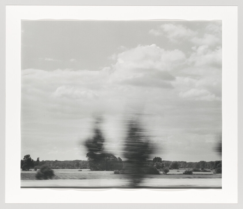 Blurred trees streak across a calm rural landscape under a cloudy sky.