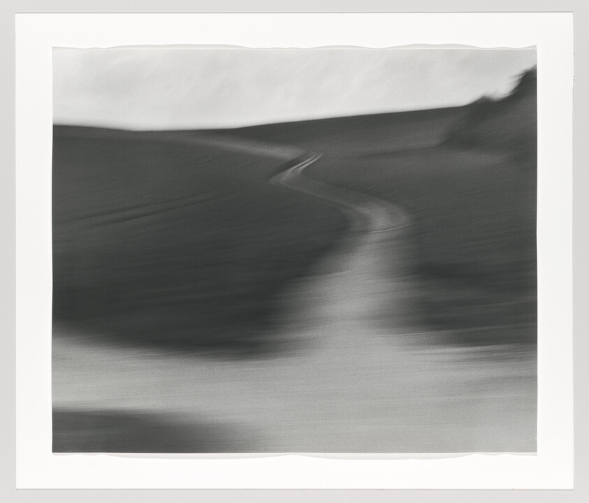 A blurred winding path curves through dark hills under a pale sky.