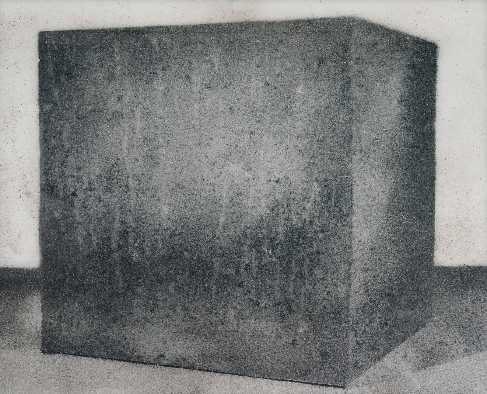 Large textured dark cube sitting on a pale floor against a light wall.