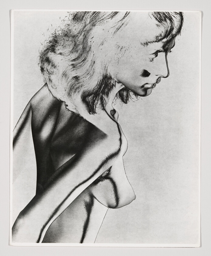 Woman turned in profile showing upper torso and bare breasts against a plain background.