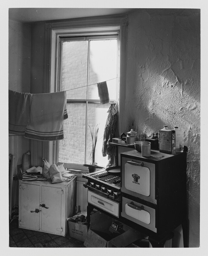 Small kitchen with an old gas stove, drying towels across the window, and a cluttered counter.