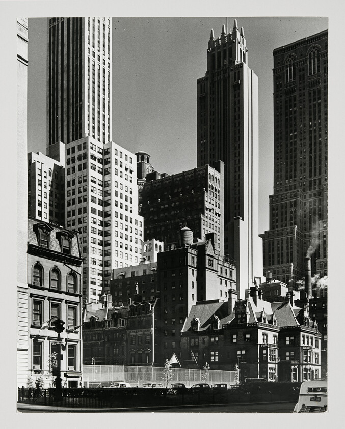 Tall skyscrapers rise behind older stone townhouses and parked cars on a city street.