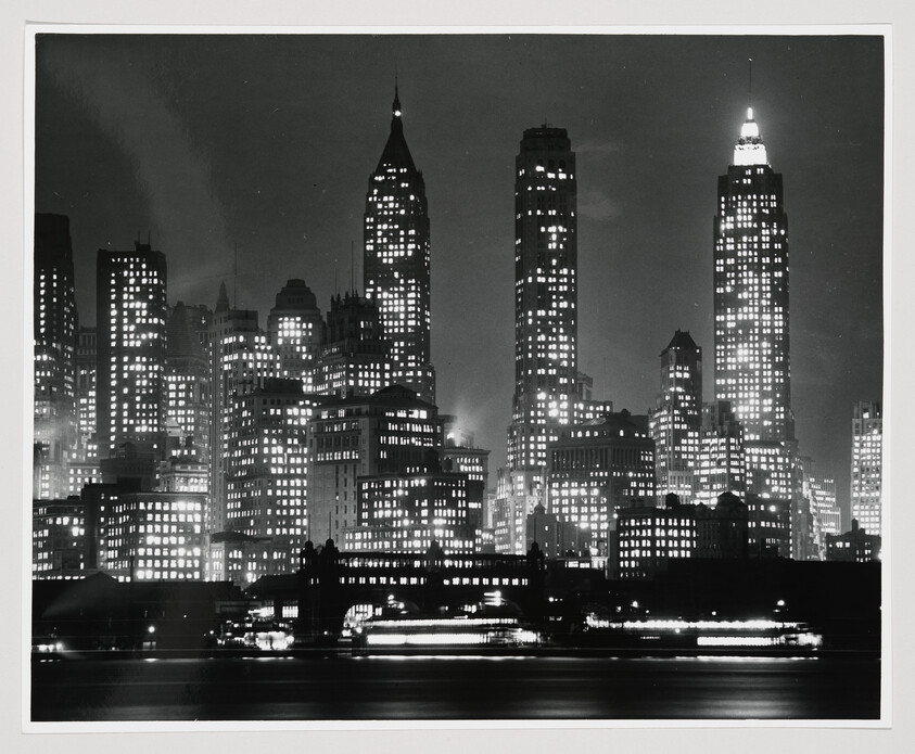 Tall illuminated skyscrapers rise above a lit waterfront with blurred boat lights at night.