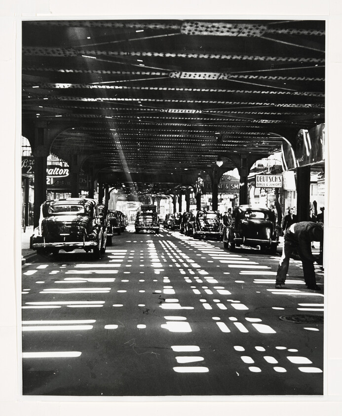 Vintage cars line a city street under elevated train tracks with strong light patterns on the pavement.