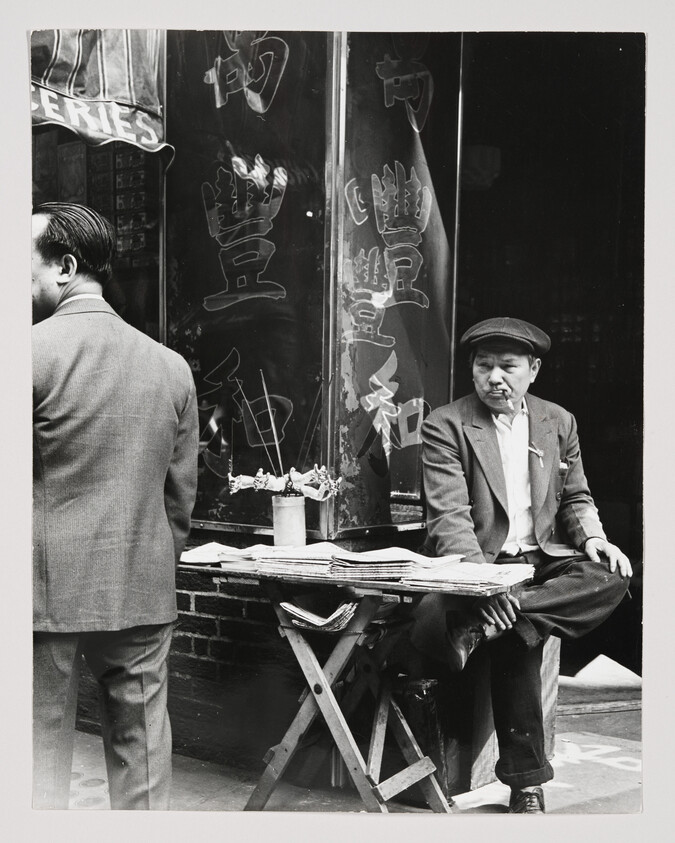 Older man sits cross-legged by a small newsstand, smoking a cigarette and selling papers.