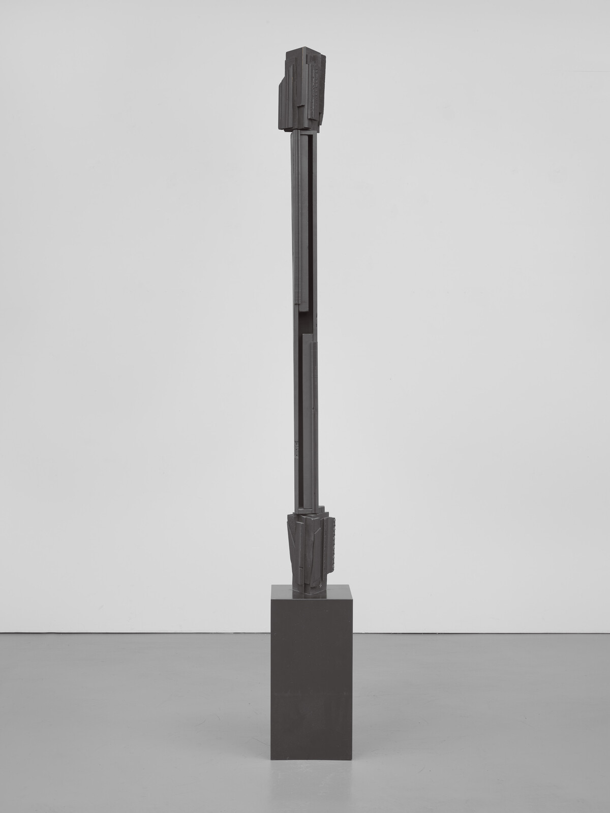 Tall, abstract metal sculpture on a square pedestal in a minimalist gallery setting.