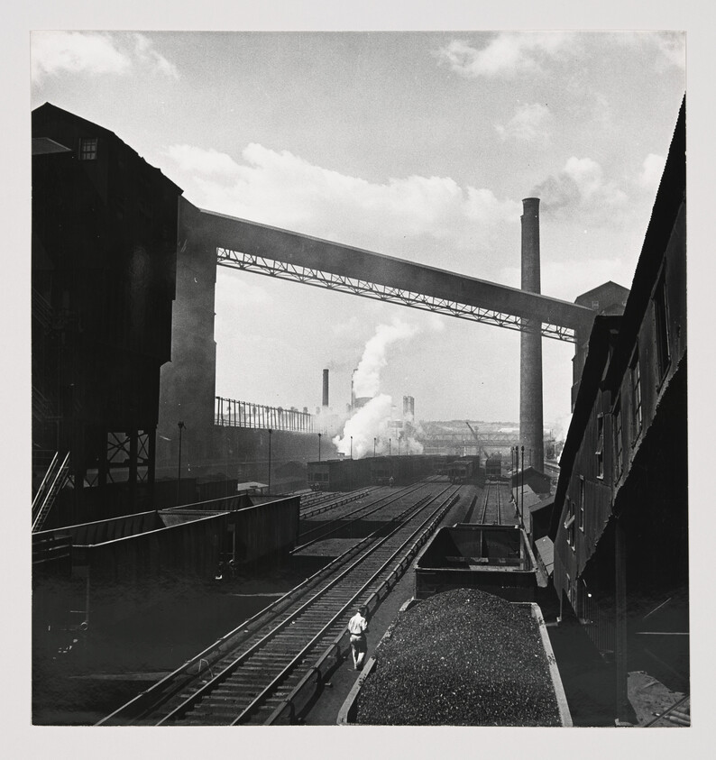 Worker walks beside coal-filled rail cars in an industrial yard with smokestacks and bridges.